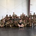 A large group of military personnel, predominantly in camouflage uniforms, pose for a group photo inside what appears to be a training facility or hangar. They are gathered around a black banner reading 'MACP' (Modern Army Combatives Program). The group includes both uniformed soldiers and a few individuals in civilian/athletic clothing, suggesting a combatives training event or course completion.