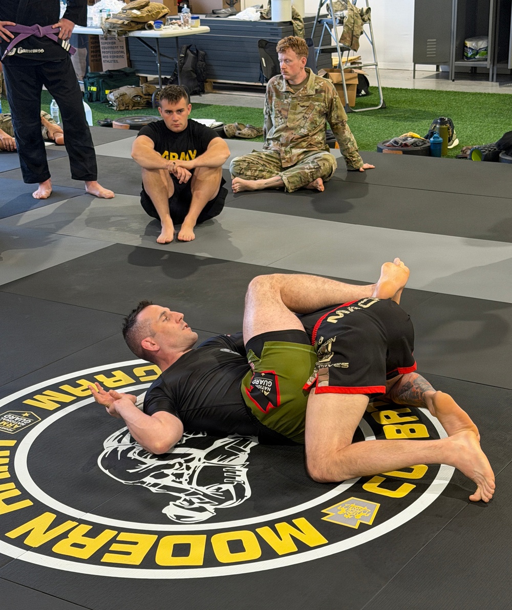 A grappling/jiu-jitsu training session is taking place on a mat bearing 'Modern Army Combatives Program (MACP)' and Army National Guard branding. Two athletes are engaged in ground fighting on the mat while several observers, including soldiers in military uniforms and civilian athletic wear, watch from the perimeter. The setting appears to be a dedicated combatives training facility.