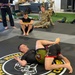 A grappling/jiu-jitsu training session is taking place on a mat bearing 'Modern Army Combatives Program (MACP)' and Army National Guard branding. Two athletes are engaged in ground fighting on the mat while several observers, including soldiers in military uniforms and civilian athletic wear, watch from the perimeter. The setting appears to be a dedicated combatives training facility.