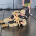 A military combatives training session is taking place on a padded mat floor. A soldier in full camouflage uniform (OCP pattern) and tan boots is grappling with a training partner wearing a black padded suit and protective helmet. A third individual in civilian athletic wear stands nearby, appearing to observe or instruct.