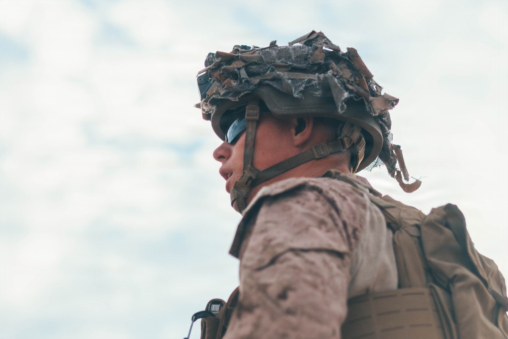 A low-angle photograph of a military service member wearing MARPAT (Marine pattern) camouflage uniform, a tactical combat helmet with camouflage netting/burlap strips attached, and blue-tinted ballistic eyewear. The subject is photographed from below against an overcast sky, giving a dramatic upward perspective. The individual appears to be equipped with a tactical backpack or plate carrier visible over the shoulder.