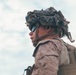 A low-angle photograph of a military service member wearing MARPAT (Marine pattern) camouflage uniform, a tactical combat helmet with camouflage netting/burlap strips attached, and blue-tinted ballistic eyewear. The subject is photographed from below against an overcast sky, giving a dramatic upward perspective. The individual appears to be equipped with a tactical backpack or plate carrier visible over the shoulder.