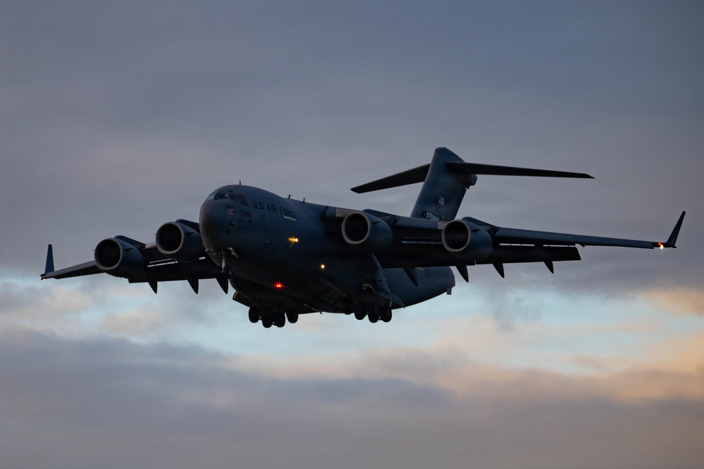 A U.S. Air Force C-17 Globemaster III military cargo aircraft is captured in flight during dusk or dawn conditions. The aircraft is photographed from a slightly low angle with landing gear deployed, suggesting it is on approach for landing. The overcast sky with muted orange and grey tones provides a dramatic backdrop.