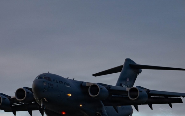 A U.S. Air Force C-17 Globemaster III military cargo aircraft is captured in flight during dusk or dawn conditions. The aircraft is photographed from a slightly low angle with landing gear deployed, suggesting it is on approach for landing. The overcast sky with muted orange and grey tones provides a dramatic backdrop.