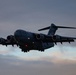 A U.S. Air Force C-17 Globemaster III military cargo aircraft is captured in flight during dusk or dawn conditions. The aircraft is photographed from a slightly low angle with landing gear deployed, suggesting it is on approach for landing. The overcast sky with muted orange and grey tones provides a dramatic backdrop.