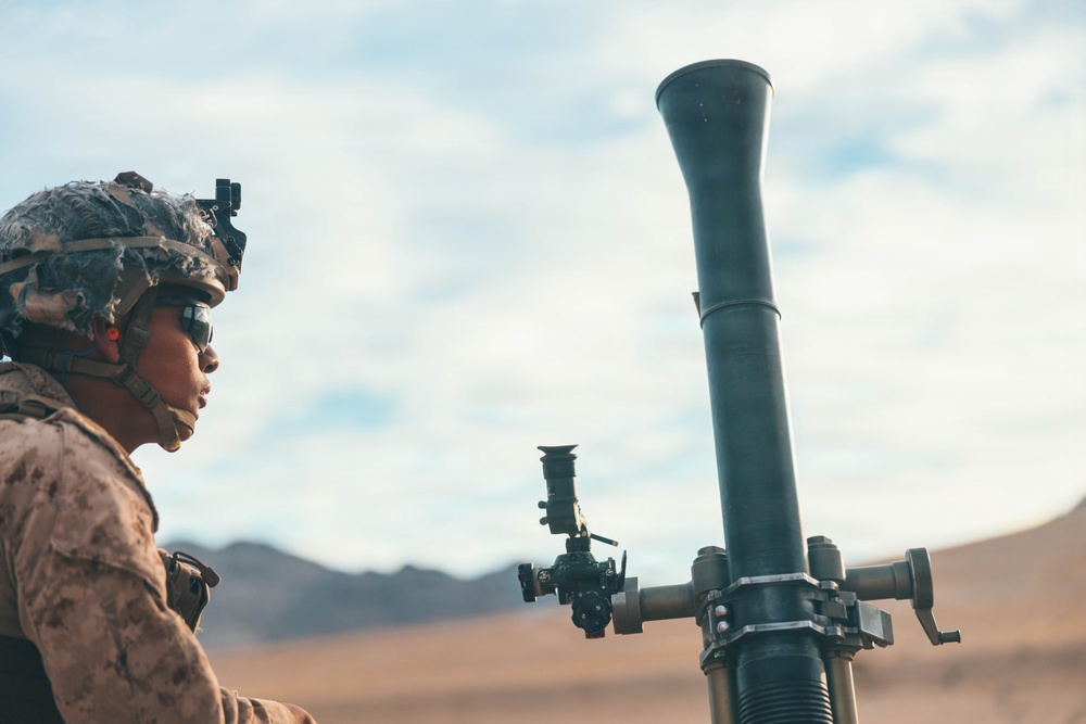 A military service member in desert camouflage uniform is positioned next to a mortar tube system in an arid, desert training environment. The soldier is wearing a combat helmet with night vision device mount and protective eyewear. The setting appears to be a live-fire training exercise based on the terrain and equipment configuration.