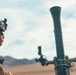 A military service member in desert camouflage uniform is positioned next to a mortar tube system in an arid, desert training environment. The soldier is wearing a combat helmet with night vision device mount and protective eyewear. The setting appears to be a live-fire training exercise based on the terrain and equipment configuration.