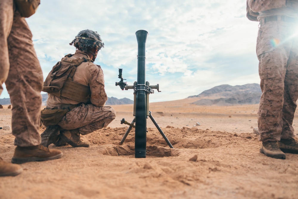 Military personnel in desert camouflage uniforms are positioned around a mortar system emplaced in sandy terrain. The mortar tube is set up in a shallow pit with its bipod and baseplate visible. The arid, mountainous landscape in the background suggests a desert training or operational environment.