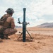 Military personnel in desert camouflage uniforms are positioned around a mortar system emplaced in sandy terrain. The mortar tube is set up in a shallow pit with its bipod and baseplate visible. The arid, mountainous landscape in the background suggests a desert training or operational environment.