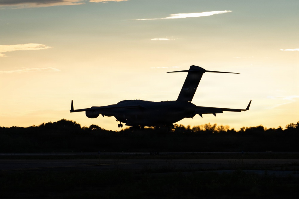 A large military or commercial aircraft is captured in silhouette during takeoff or landing at dusk, with a warm golden sunset sky in the background. The aircraft appears to be a large transport or cargo-type plane based on its high-wing configuration and T-tail design. The low-light conditions create a dramatic contrast between the dark aircraft silhouette and the illuminated sky.