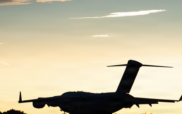 A large military or commercial aircraft is captured in silhouette during takeoff or landing at dusk, with a warm golden sunset sky in the background. The aircraft appears to be a large transport or cargo-type plane based on its high-wing configuration and T-tail design. The low-light conditions create a dramatic contrast between the dark aircraft silhouette and the illuminated sky.