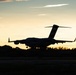 A large military or commercial aircraft is captured in silhouette during takeoff or landing at dusk, with a warm golden sunset sky in the background. The aircraft appears to be a large transport or cargo-type plane based on its high-wing configuration and T-tail design. The low-light conditions create a dramatic contrast between the dark aircraft silhouette and the illuminated sky.