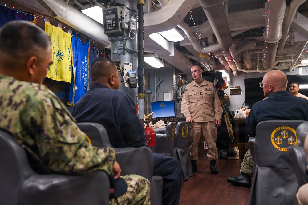 A U.S. Navy briefing or meeting is taking place in what appears to be a Chief Petty Officers' mess aboard a naval vessel. A standing officer in tan/khaki uniform addresses seated personnel wearing various Navy uniforms including camouflage and working blues. The space is decorated with holiday lights and displays Navy flags and unit banners.