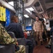 A U.S. Navy briefing or meeting is taking place in what appears to be a Chief Petty Officers' mess aboard a naval vessel. A standing officer in tan/khaki uniform addresses seated personnel wearing various Navy uniforms including camouflage and working blues. The space is decorated with holiday lights and displays Navy flags and unit banners.
