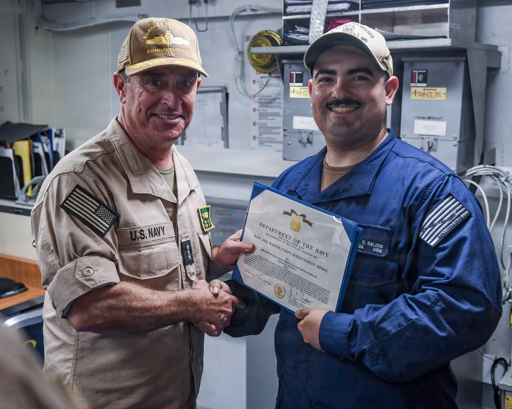 Two U.S. Navy personnel are shaking hands during a military award ceremony aboard what appears to be a naval vessel. The sailor on the right, identified by his name tag as 'R. Salido,' is receiving a Navy and Marine Corps Achievement Medal certificate from a senior officer wearing a 'COMNAVSURFPAC' cap. Both individuals are smiling, indicating a positive and celebratory occasion.