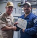 Two U.S. Navy personnel are shaking hands during a military award ceremony aboard what appears to be a naval vessel. The sailor on the right, identified by his name tag as 'R. Salido,' is receiving a Navy and Marine Corps Achievement Medal certificate from a senior officer wearing a 'COMNAVSURFPAC' cap. Both individuals are smiling, indicating a positive and celebratory occasion.