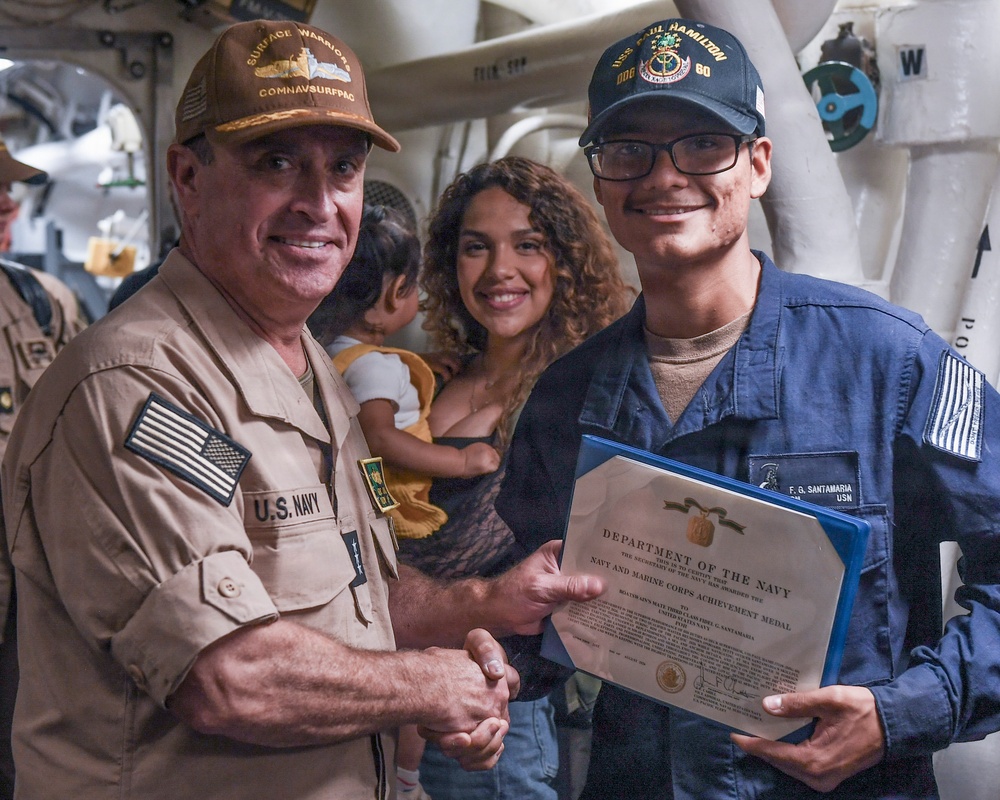 A U.S. Navy award ceremony taking place aboard a naval vessel, where a senior officer is presenting the Navy and Marine Corps Achievement Medal to a sailor named F.G. Santamaria. The recipient is holding a Department of the Navy certificate in a blue folder while shaking hands with the presenting officer. A woman holding a young child stands in the background, likely a family member of the recipient.
