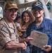 A U.S. Navy award ceremony taking place aboard a naval vessel, where a senior officer is presenting the Navy and Marine Corps Achievement Medal to a sailor named F.G. Santamaria. The recipient is holding a Department of the Navy certificate in a blue folder while shaking hands with the presenting officer. A woman holding a young child stands in the background, likely a family member of the recipient.