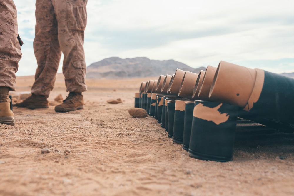 The image shows military personnel in desert camouflage uniforms standing beside a row of expended artillery or mortar shell casings laid out on arid desert ground. The casings appear to be black metal with tan/cardboard propellant charge containers, arranged in a line. The setting is a desert training or firing range environment with mountains visible in the background.
