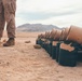 The image shows military personnel in desert camouflage uniforms standing beside a row of expended artillery or mortar shell casings laid out on arid desert ground. The casings appear to be black metal with tan/cardboard propellant charge containers, arranged in a line. The setting is a desert training or firing range environment with mountains visible in the background.