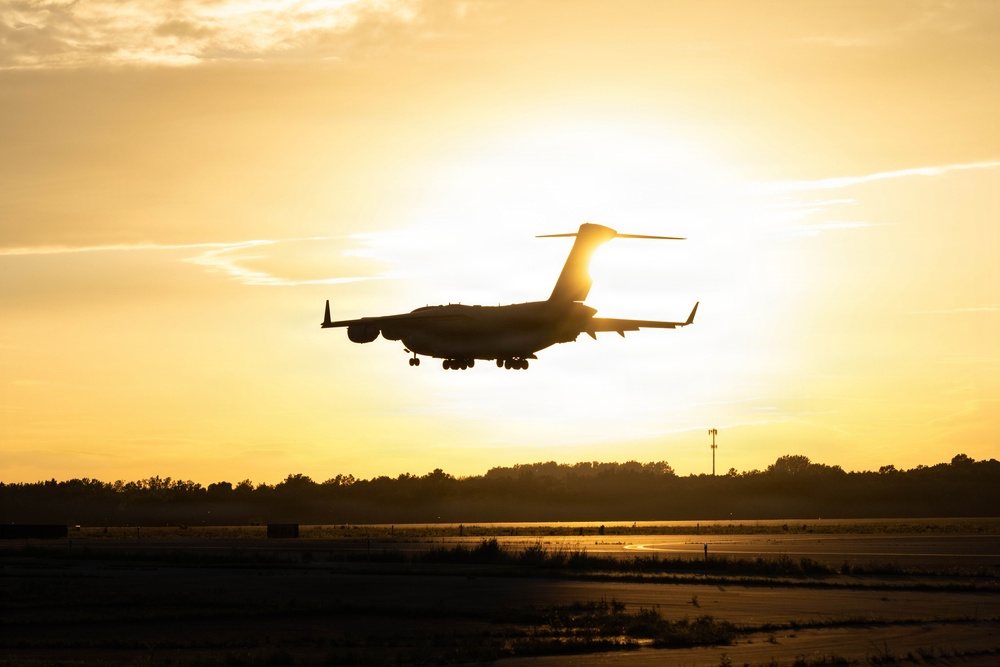 A large military transport aircraft, consistent with a C-17 Globemaster III silhouette, is captured on final approach to land at an airfield during golden hour or sunset. The aircraft is backlit by the sun, creating a dramatic silhouette effect against a warm yellow-orange sky. Landing gear is visibly deployed, indicating imminent touchdown on the runway below.