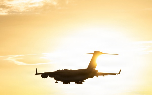 A large military transport aircraft, consistent with a C-17 Globemaster III silhouette, is captured on final approach to land at an airfield during golden hour or sunset. The aircraft is backlit by the sun, creating a dramatic silhouette effect against a warm yellow-orange sky. Landing gear is visibly deployed, indicating imminent touchdown on the runway below.