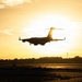 A large military transport aircraft, consistent with a C-17 Globemaster III silhouette, is captured on final approach to land at an airfield during golden hour or sunset. The aircraft is backlit by the sun, creating a dramatic silhouette effect against a warm yellow-orange sky. Landing gear is visibly deployed, indicating imminent touchdown on the runway below.