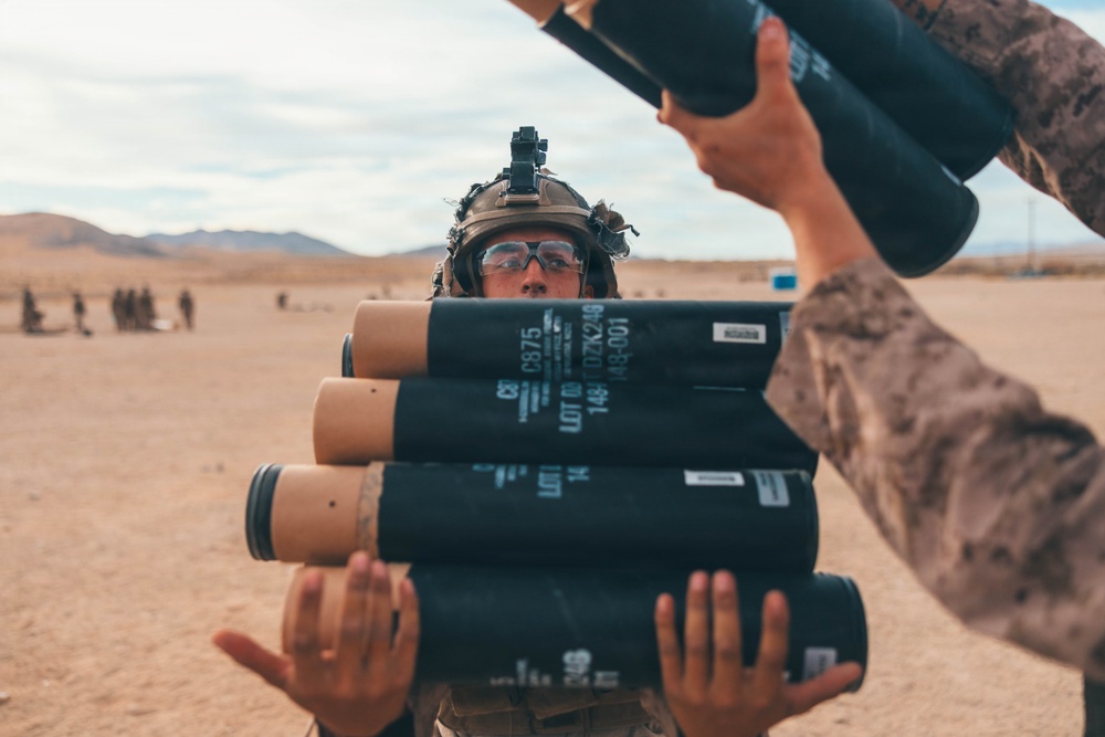 A military service member in desert camouflage uniform and combat helmet with night vision mount is carrying multiple cylindrical munition containers in an arid, desert training environment. Another service member's arms are visible passing additional containers. The setting appears to be a live-fire training exercise based on the desert terrain and presence of other personnel in the background.