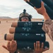 A military service member in desert camouflage uniform and combat helmet with night vision mount is carrying multiple cylindrical munition containers in an arid, desert training environment. Another service member's arms are visible passing additional containers. The setting appears to be a live-fire training exercise based on the desert terrain and presence of other personnel in the background.