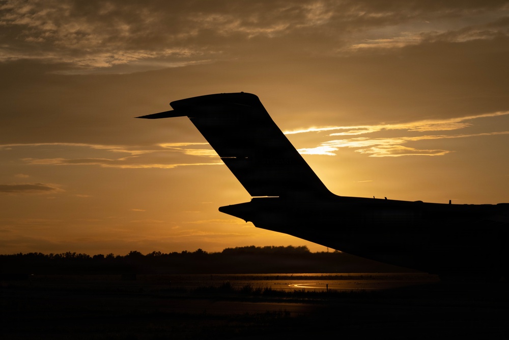 A dramatic silhouette photograph of an aircraft tail section (empennage) captured against a golden sunset sky. The image is taken from ground level on what appears to be an airport tarmac, with the sun positioned directly behind the tail structure. The warm amber and orange tones of the sunset create a striking contrast against the dark aircraft silhouette.