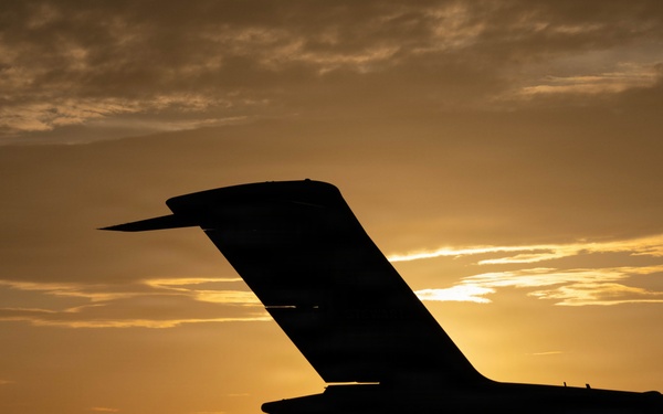 A dramatic silhouette photograph of an aircraft tail section (empennage) captured against a golden sunset sky. The image is taken from ground level on what appears to be an airport tarmac, with the sun positioned directly behind the tail structure. The warm amber and orange tones of the sunset create a striking contrast against the dark aircraft silhouette.