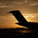 A dramatic silhouette photograph of an aircraft tail section (empennage) captured against a golden sunset sky. The image is taken from ground level on what appears to be an airport tarmac, with the sun positioned directly behind the tail structure. The warm amber and orange tones of the sunset create a striking contrast against the dark aircraft silhouette.