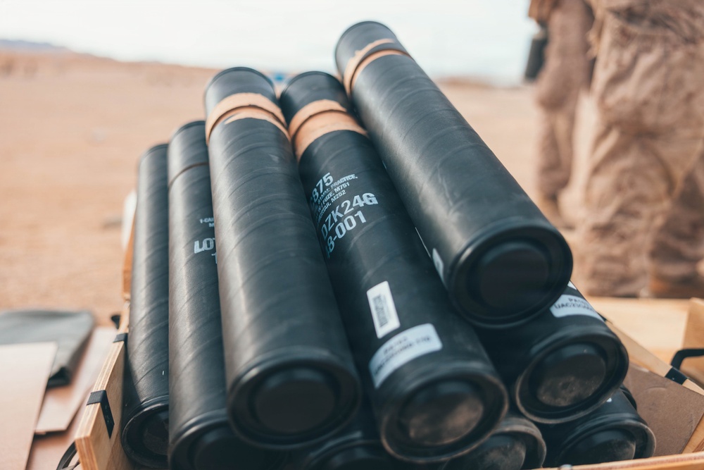 The image shows a stack of black cylindrical munition containers stored in a wooden crate in an arid, desert environment. The containers display visible lot and identification markings. A person in desert camouflage uniform is partially visible in the background.