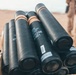 The image shows a stack of black cylindrical munition containers stored in a wooden crate in an arid, desert environment. The containers display visible lot and identification markings. A person in desert camouflage uniform is partially visible in the background.