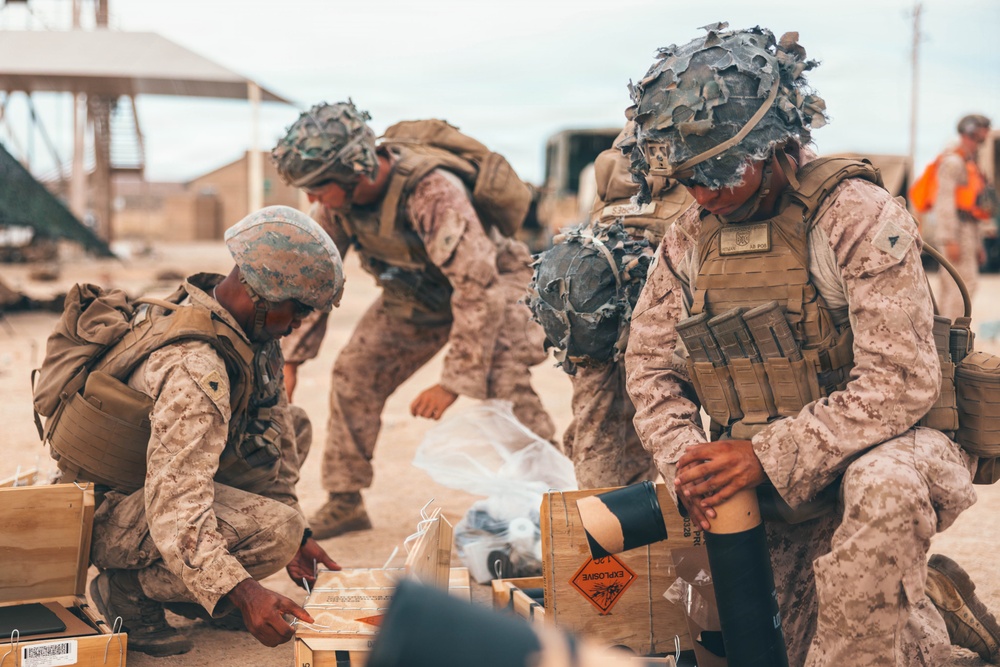 Three military personnel in MARPAT desert camouflage uniforms are handling munitions and equipment from wooden crates in what appears to be a desert training environment. The soldiers are wearing tactical vests, helmets with camouflage netting, and face paint, suggesting a field exercise or operational scenario. A wooden crate with an 'EXPLOSIVE' hazard label is visible in the foreground.