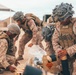 Three military personnel in MARPAT desert camouflage uniforms are handling munitions and equipment from wooden crates in what appears to be a desert training environment. The soldiers are wearing tactical vests, helmets with camouflage netting, and face paint, suggesting a field exercise or operational scenario. A wooden crate with an 'EXPLOSIVE' hazard label is visible in the foreground.