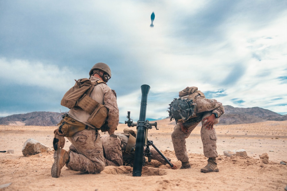Two military personnel in desert camouflage uniforms are operating a mortar system in an arid, desert training environment. A mortar round is visibly captured mid-flight above the tube, indicating the weapon has just been fired. The setting appears to be a military training exercise based on the controlled environment and desert terrain.