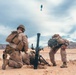 Two military personnel in desert camouflage uniforms are operating a mortar system in an arid, desert training environment. A mortar round is visibly captured mid-flight above the tube, indicating the weapon has just been fired. The setting appears to be a military training exercise based on the controlled environment and desert terrain.