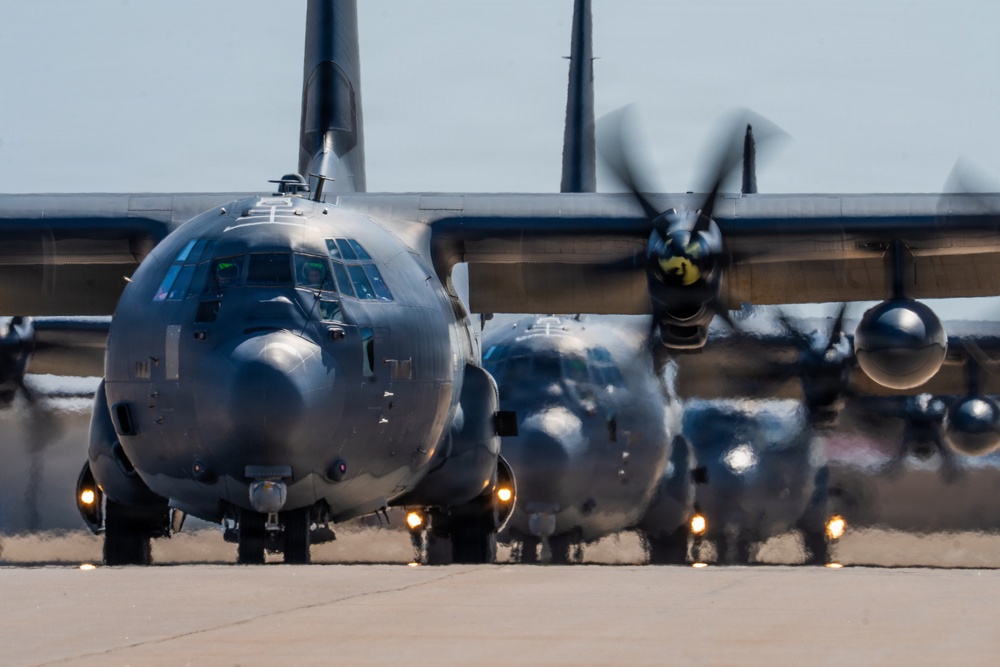 A formation of military turboprop aircraft, consistent with C-130 Hercules variants, are taxiing in a line on a runway or taxiway. The lead aircraft is sharply in focus while heat distortion from engine exhaust blurs the trailing aircraft. Propellers on the nearest aircraft are visibly spinning, indicating active engine operation.