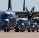 A formation of military turboprop aircraft, consistent with C-130 Hercules variants, are taxiing in a line on a runway or taxiway. The lead aircraft is sharply in focus while heat distortion from engine exhaust blurs the trailing aircraft. Propellers on the nearest aircraft are visibly spinning, indicating active engine operation.
