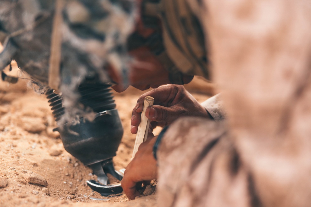 The image shows a person in camouflage or desert-patterned clothing working close to the ground in a sandy, arid environment. They appear to be handling a small rolled piece of material, possibly bark or paper, near a dark smoking object on the ground. The scene suggests a traditional fire-starting or craft activity in a desert setting.