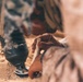 The image shows a person in camouflage or desert-patterned clothing working close to the ground in a sandy, arid environment. They appear to be handling a small rolled piece of material, possibly bark or paper, near a dark smoking object on the ground. The scene suggests a traditional fire-starting or craft activity in a desert setting.