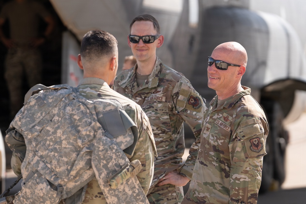 Three military personnel are engaged in conversation on what appears to be a flight line or airfield tarmac. Two individuals wearing US Air Force OCP (Operational Camouflage Pattern) uniforms are facing a third person in US Army ACU (Army Combat Uniform) pattern. A military aircraft is partially visible in the background.