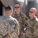 Three military personnel are engaged in conversation on what appears to be a flight line or airfield tarmac. Two individuals wearing US Air Force OCP (Operational Camouflage Pattern) uniforms are facing a third person in US Army ACU (Army Combat Uniform) pattern. A military aircraft is partially visible in the background.