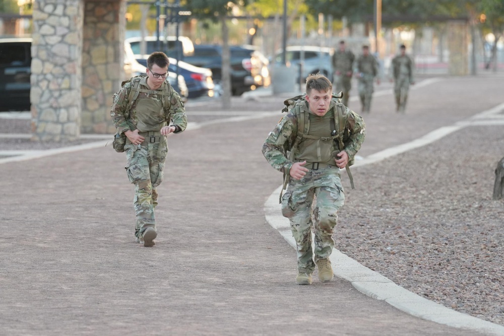 Two soldiers in US Army camouflage (OCP/MultiCam) uniforms are running or rucking along a paved pathway while carrying backpacks/rucksacks. Additional soldiers are visible in the background performing the same activity. The setting appears to be a military installation with palm trees, stone pillars, and a parking area visible.