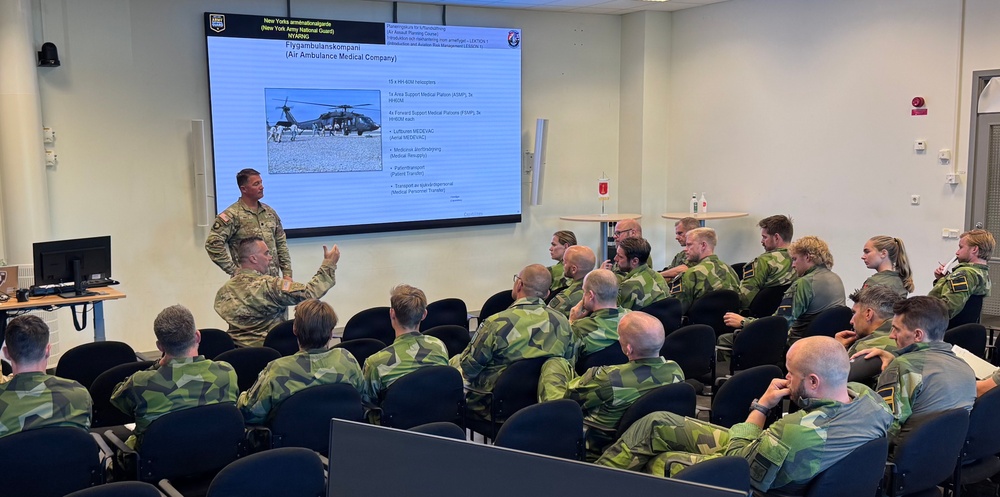 Two U.S. Army soldiers in MultiCam/OCP uniforms are presenting a military briefing to a room full of Swedish Armed Forces personnel wearing M90 camouflage uniforms. The presentation displayed on a large screen is titled 'Flygambulanskompani (Air Ambulance Medical Company)' and includes information about the New York Army National Guard (NYARNG). The setting appears to be a formal military classroom or conference room, suggesting a bilateral training or exchange program between U.S. and Swedish military forces.