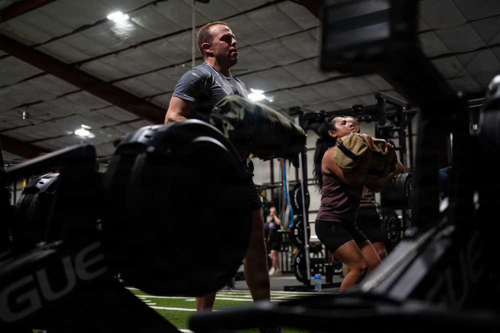 Two athletes are training in a gym facility, each carrying heavy sandbags. A male athlete in a gray shirt is in the foreground holding a large dark sandbag, while a female athlete in a maroon top carries a tan/olive sandbag behind him. The setting appears to be a functional fitness or CrossFit-style gym with turf flooring and Rogue equipment visible.