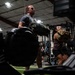 Two athletes are training in a gym facility, each carrying heavy sandbags. A male athlete in a gray shirt is in the foreground holding a large dark sandbag, while a female athlete in a maroon top carries a tan/olive sandbag behind him. The setting appears to be a functional fitness or CrossFit-style gym with turf flooring and Rogue equipment visible.