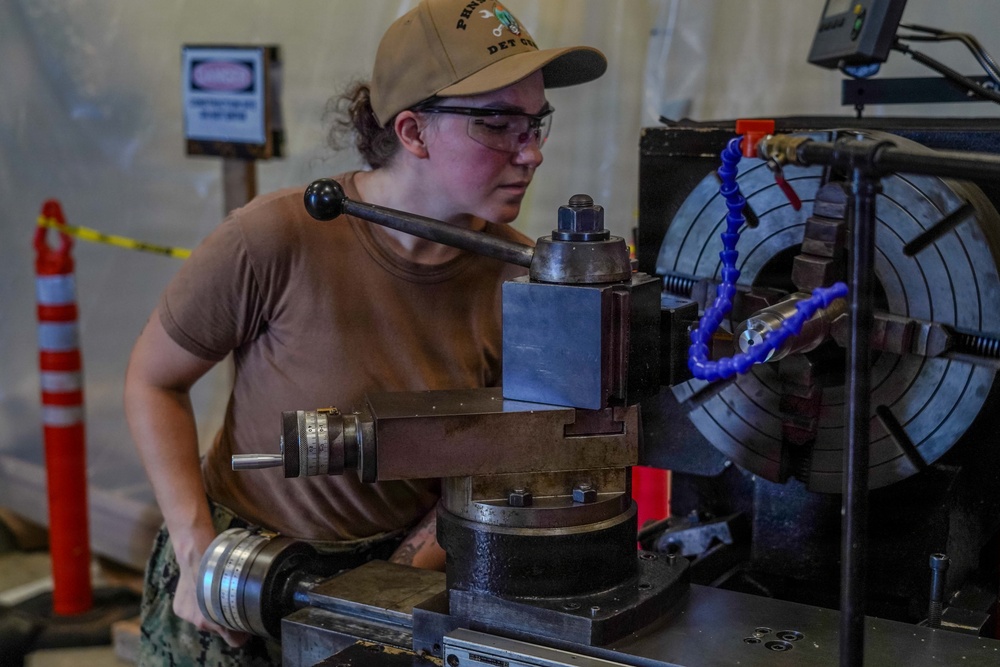 A woman wearing a tan baseball cap, safety glasses, and a brown t-shirt is operating a metal lathe in what appears to be a workshop or machine shop. She is focused on manipulating the lathe's controls, with a large chuck visible on the right side of the machine. The setting includes safety signage and traffic cones visible in the background, suggesting an industrial or military maintenance environment.