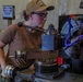 A woman wearing a tan baseball cap, safety glasses, and a brown t-shirt is operating a metal lathe in what appears to be a workshop or machine shop. She is focused on manipulating the lathe's controls, with a large chuck visible on the right side of the machine. The setting includes safety signage and traffic cones visible in the background, suggesting an industrial or military maintenance environment.
